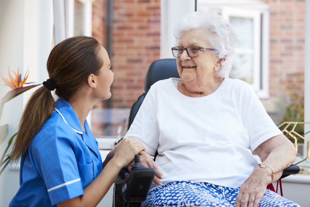 Senior Woman Sitting In Motorized Wheelchair Talking With Nurse In Retirement Home