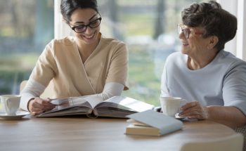 Happy senior woman spending time together with a tender caregiver sitting by a table and looking at family photo album against blurred background of windows
