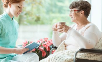 Assistant in blue uniform reading a book to elder woman in wheelchair in a hospital room
