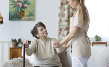 Young nurse helping sad elderly woman to stand up from the sofa